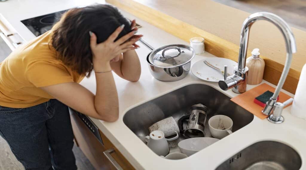 Stressed Young Woman Touching Head While Looking At Pile Of Dirty Dishes