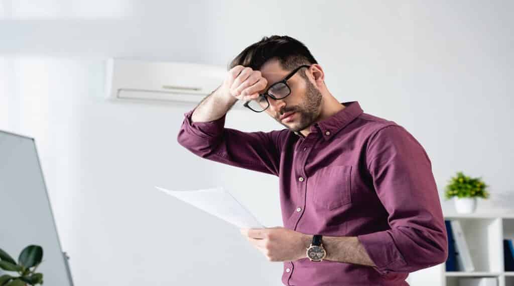 young businessman looking at document and touching forehead while suffering from heat in office